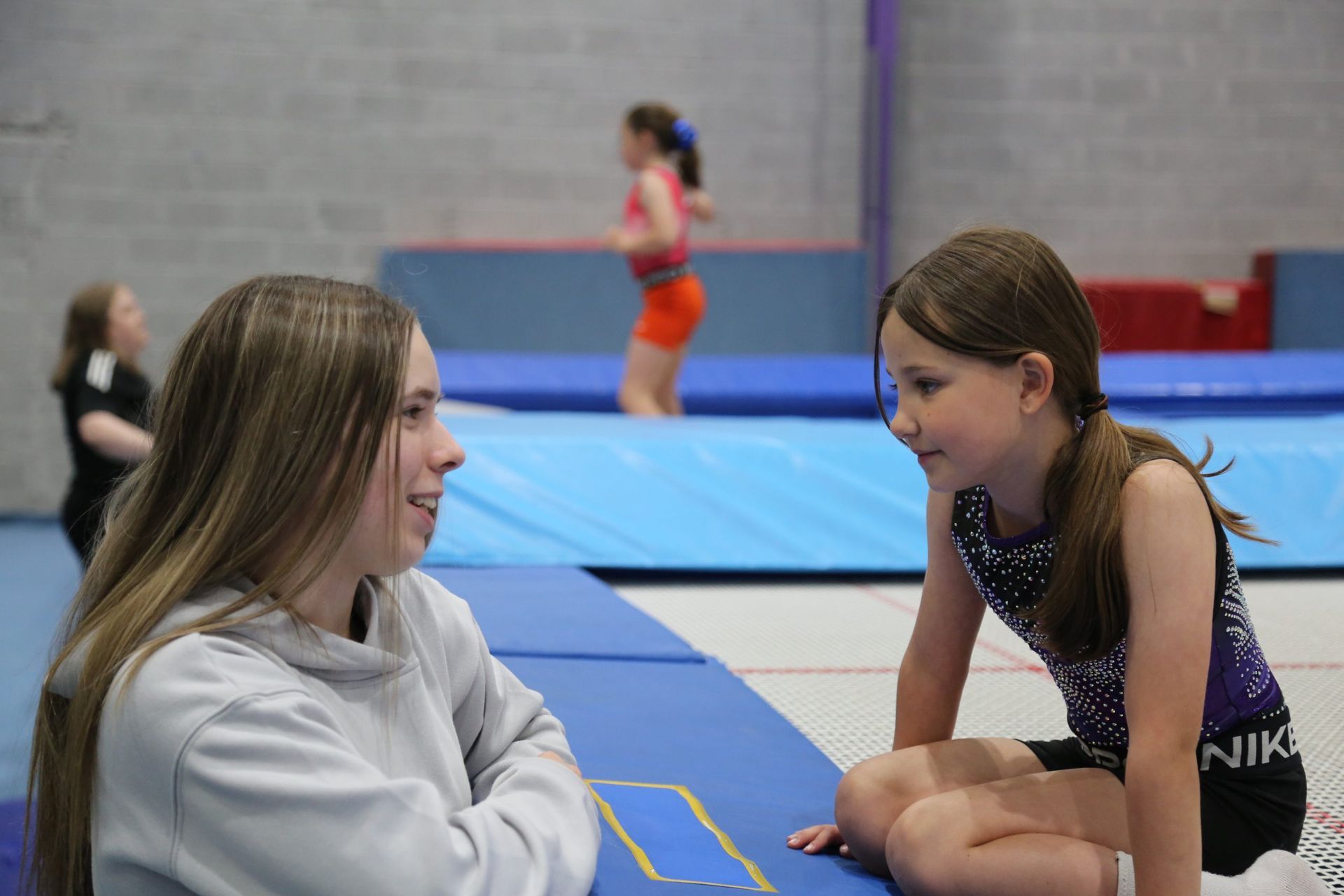 Two girls talking near a trampoline in a gym; others in the background practice.