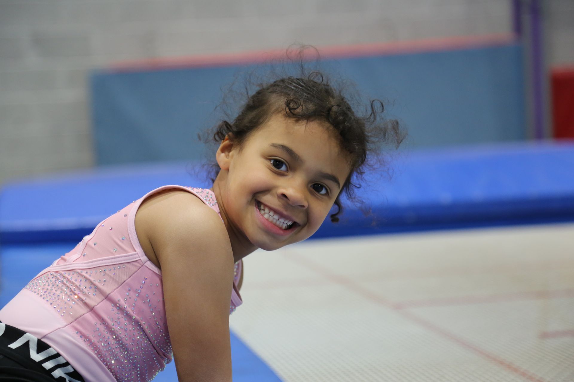 Girl smiling at the camera in a gym setting, wearing a pink top, and black shorts.