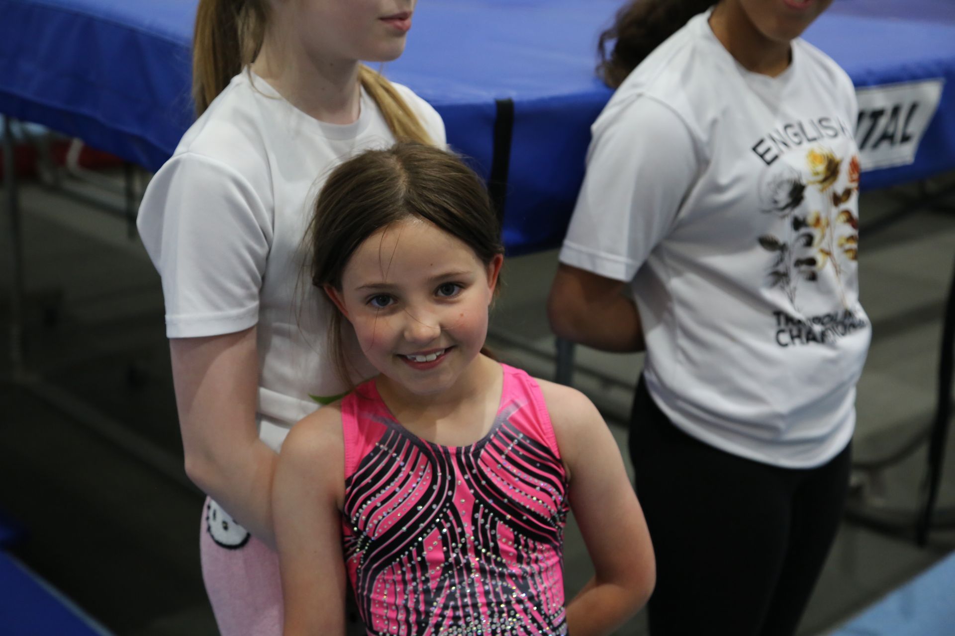 Girl in pink leotard smiles, flanked by two others in white shirts, near a blue trampoline.