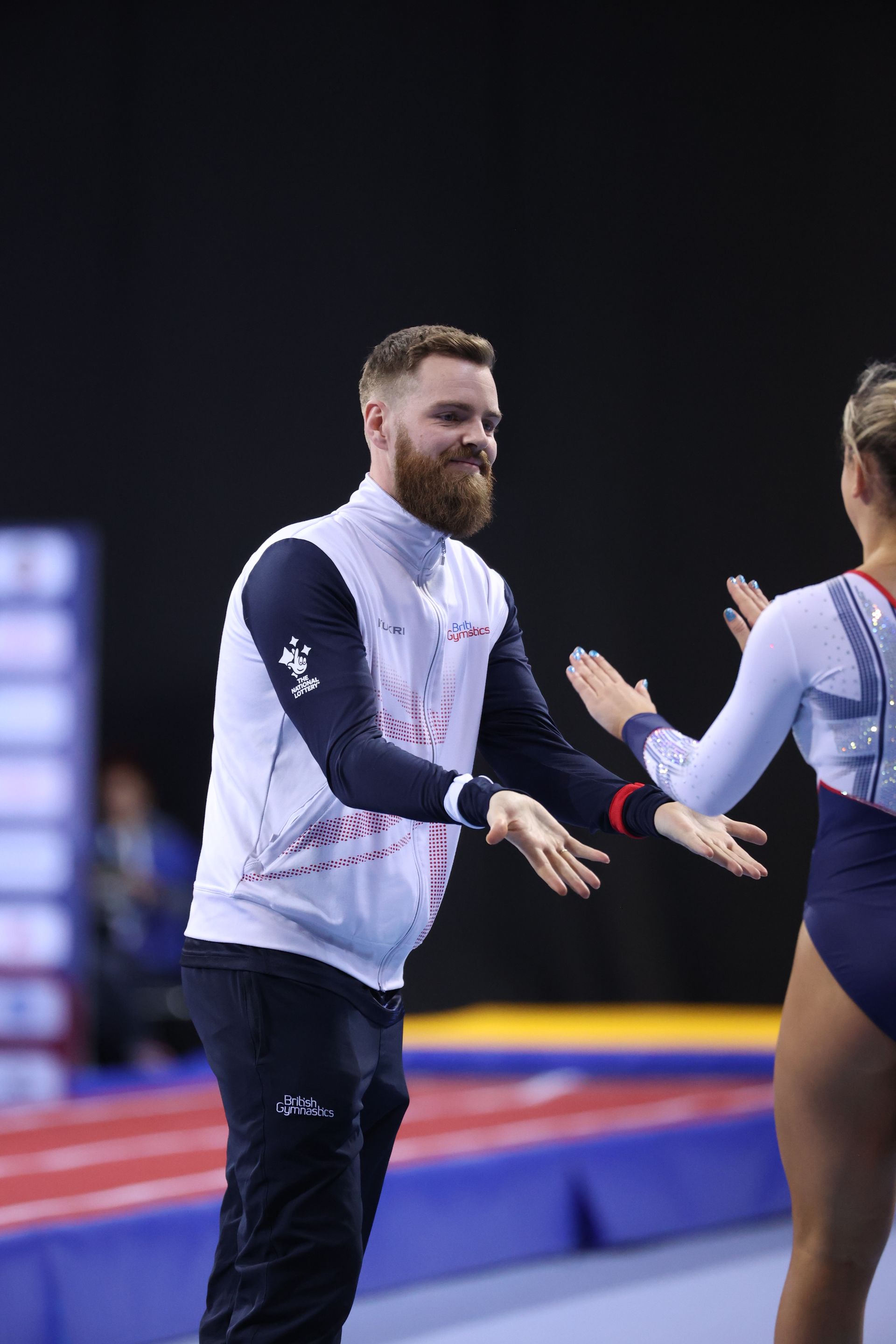 Man in white vest high-fives a gymnast, on a blue mat.