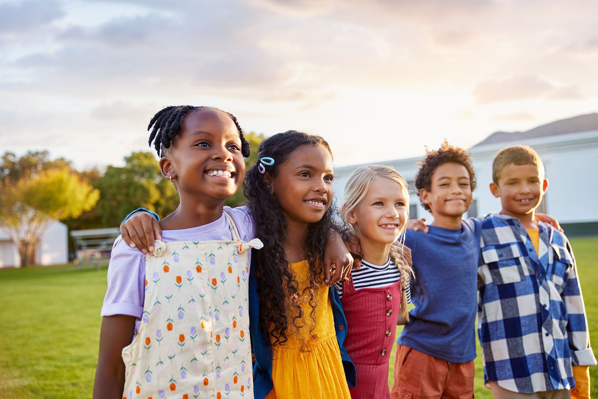 Children with arms around each other, smiling outdoors. Green lawn, sunny sky, and building in background.