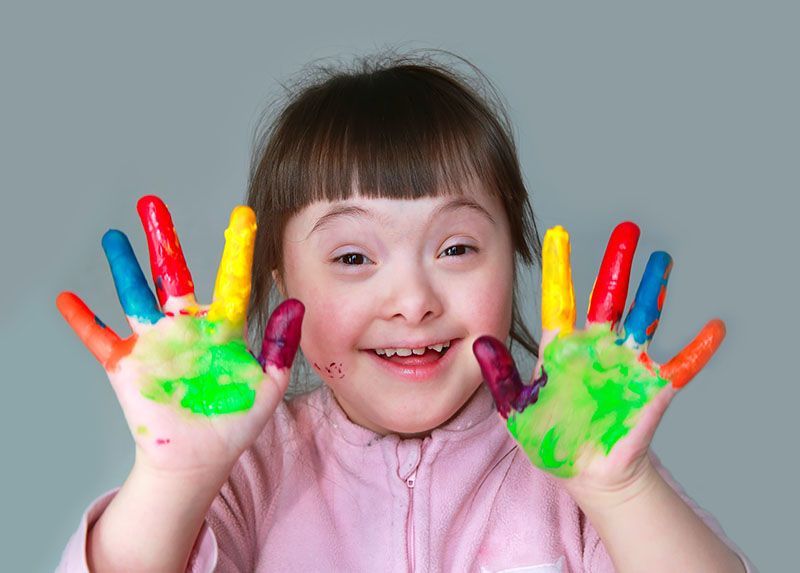 Girl with Down syndrome smiles, holding up hands covered in colorful paint.