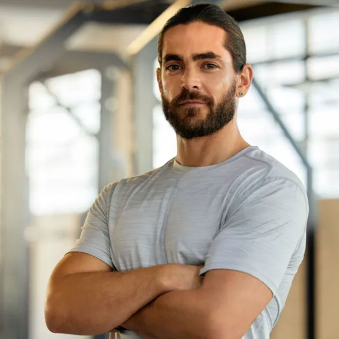 Man with arms crossed, in a gym, wearing a light gray t-shirt. He has a beard and is looking at the camera.