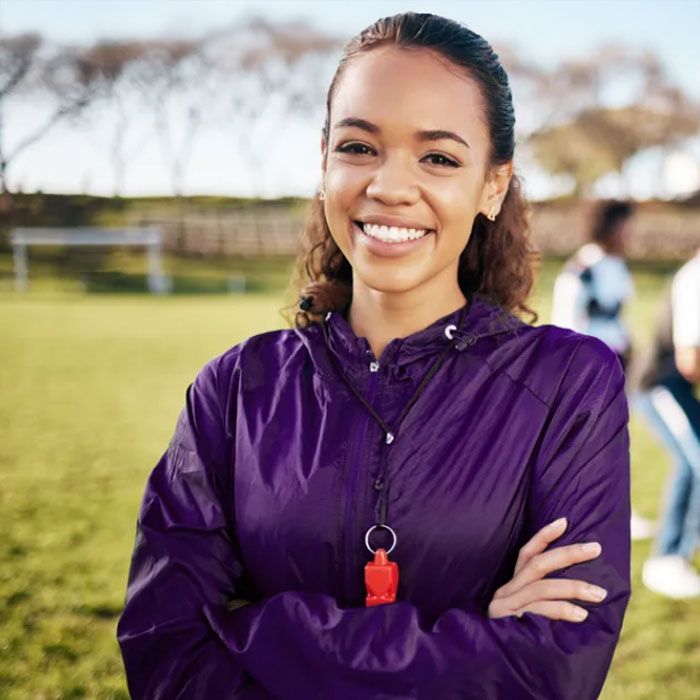 Woman in purple jacket with whistle smiles on a sports field, arms crossed.
