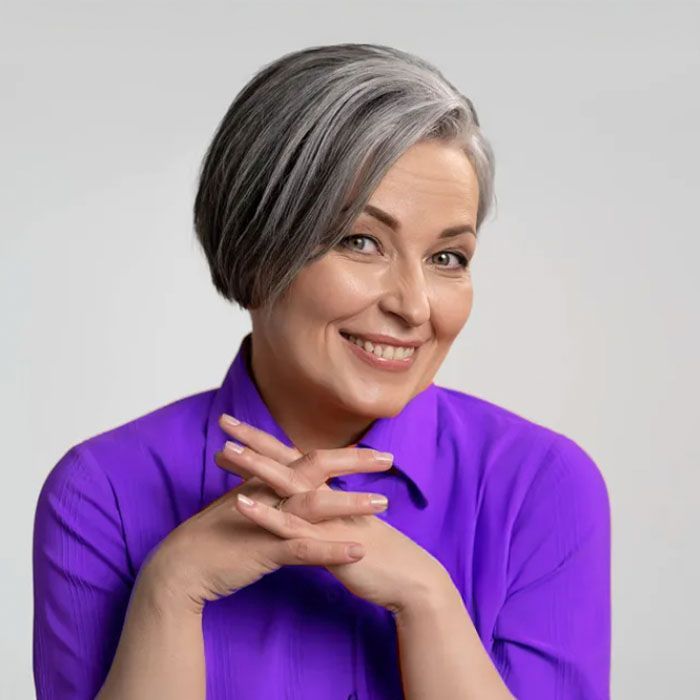 Woman with short gray hair, wearing a purple shirt, smiling, hands clasped.
