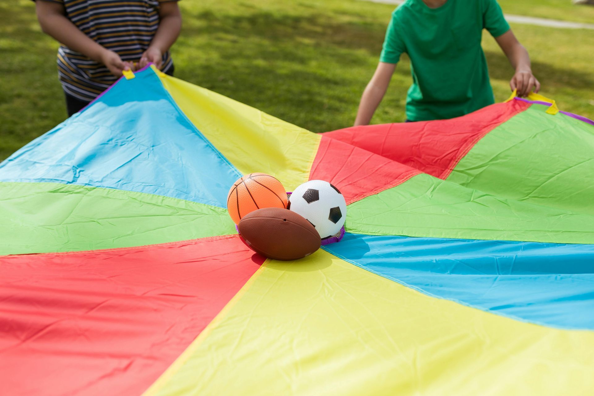 Children holding a colorful parachute, with a basketball, soccer ball, and football inside on a grassy field.