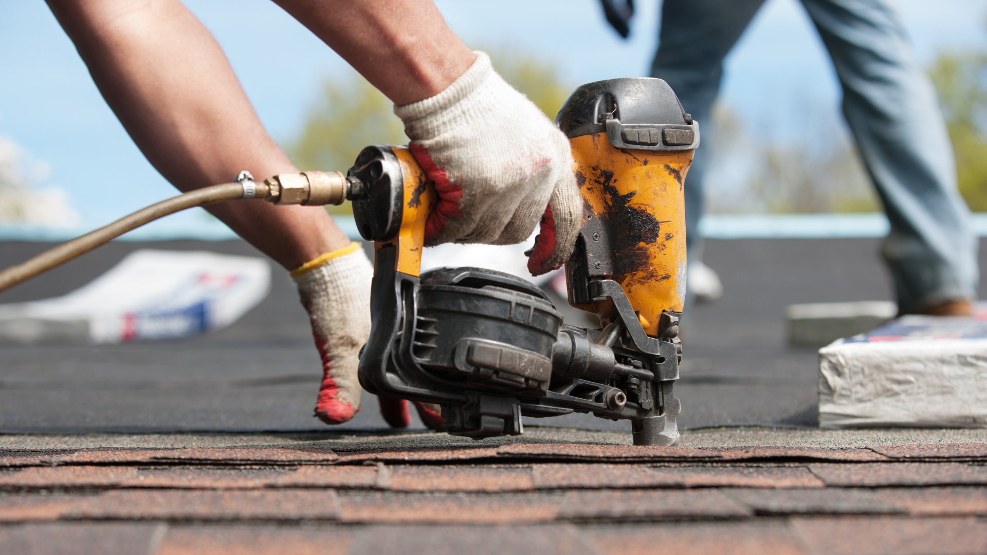 Gloved hands use a nail gun to fasten roofing shingles on a roof