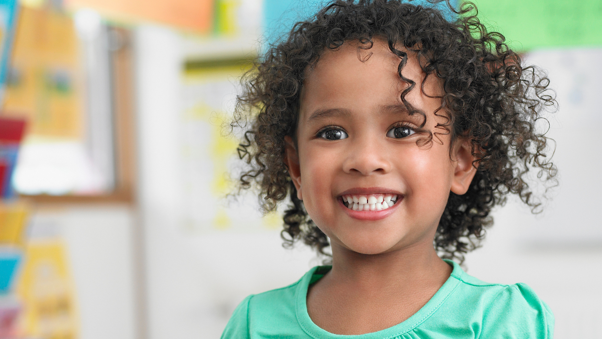 A little girl with curly hair is smiling in a classroom.