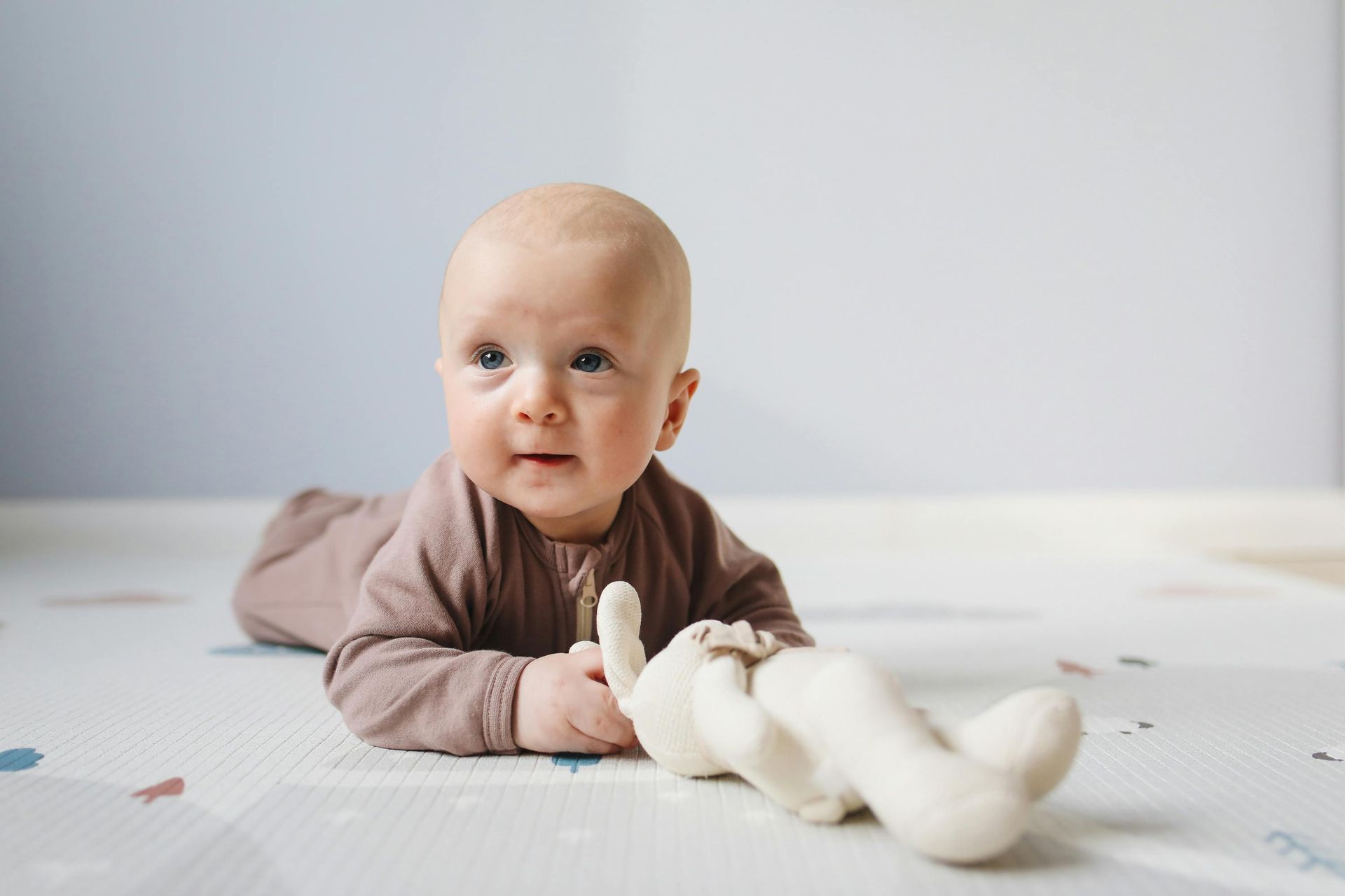 A baby is laying on its stomach on a mat holding a teddy bear.