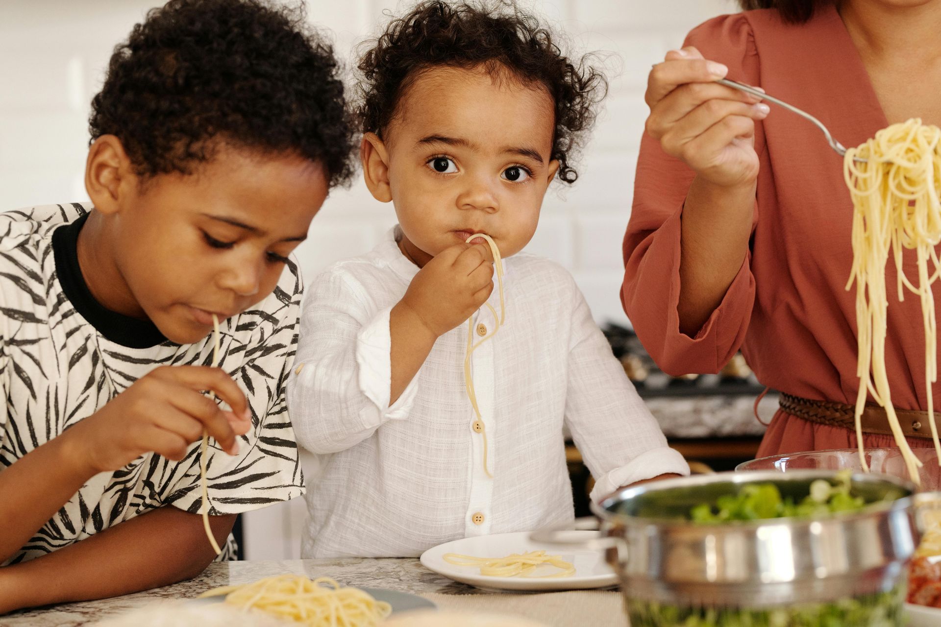 A woman and two children are eating spaghetti together in a kitchen.