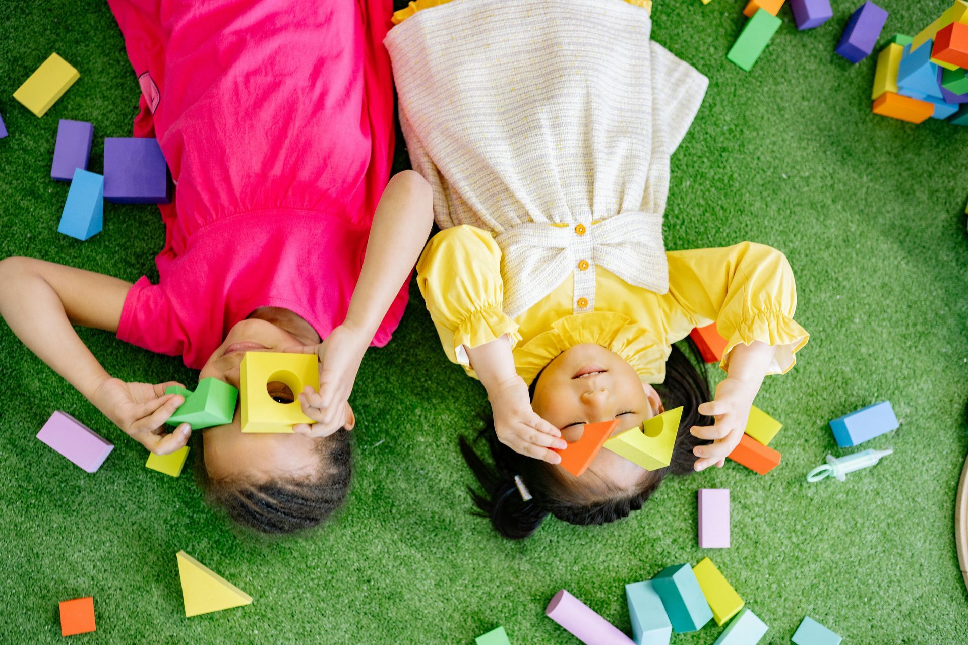 A boy and a girl are laying upside down on the grass playing with blocks.