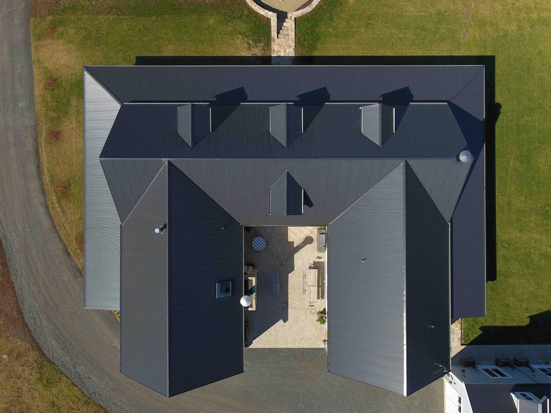 Image is of a satellite perspective directly over the house. It highlights the entire Colorbond roof, dark in colour  and almost in the shape of the capital letter H. It boasts multiple pitches which emphasises the versatility of the Colorbond product. The home is surrounded by kept green lawns and gravel driveway.  