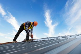 Image depicts a metal roof taken from the gutter up towards the sky. There is a man dressed in trade clothing including a hard hat and gloves. He is bending over and working on the roof as he appears to be using a drill to fix screws to the roof.