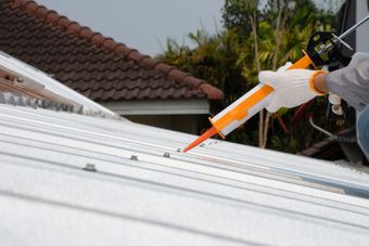 Image depicts a metal roof with close ups of tex screws fixing the metal down. There is a trades person coming into the shot holding a silicon gun administering silicon to seal around the screws.