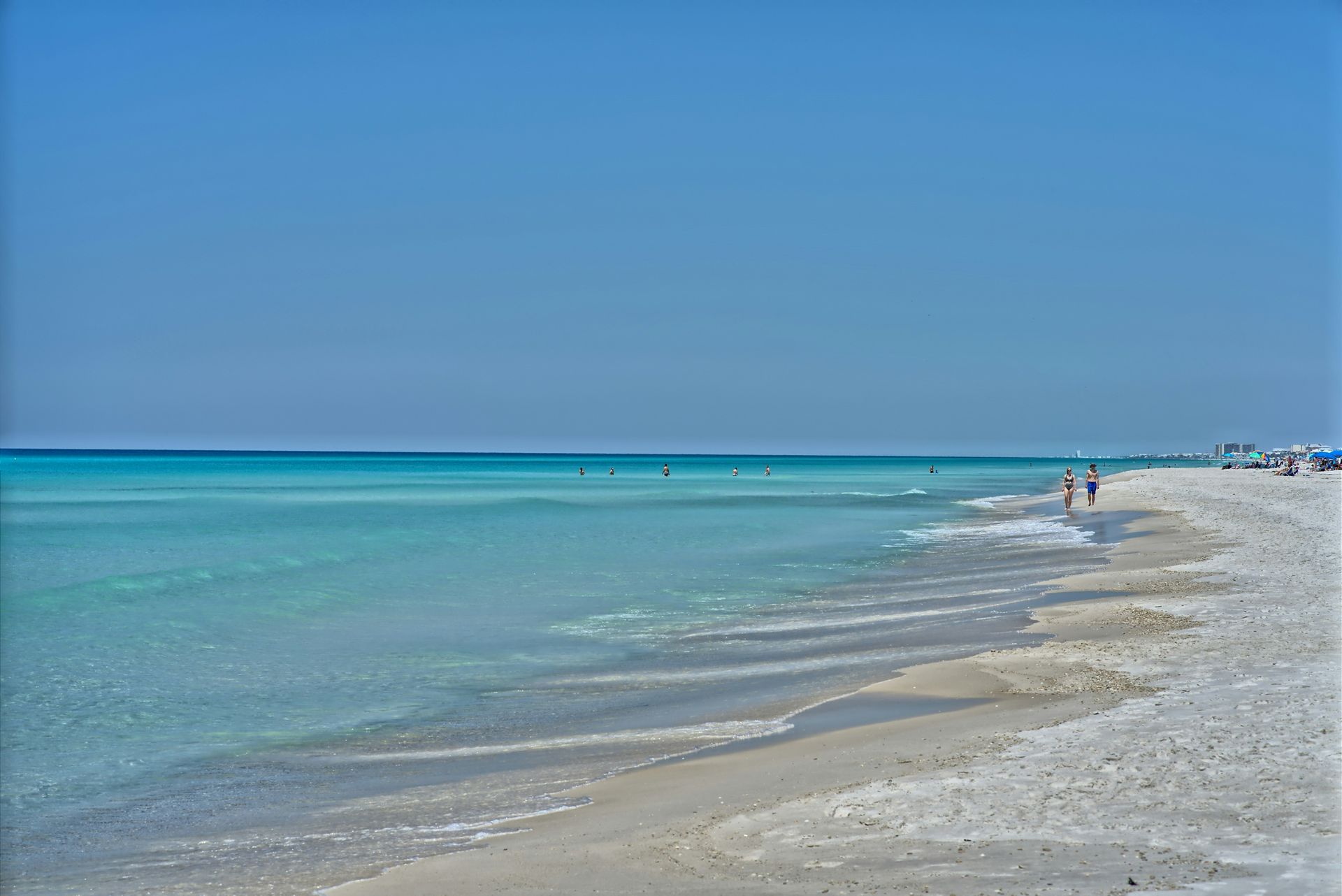 Beach with turquoise water, white sand, and clear blue sky; people walking along the shore.