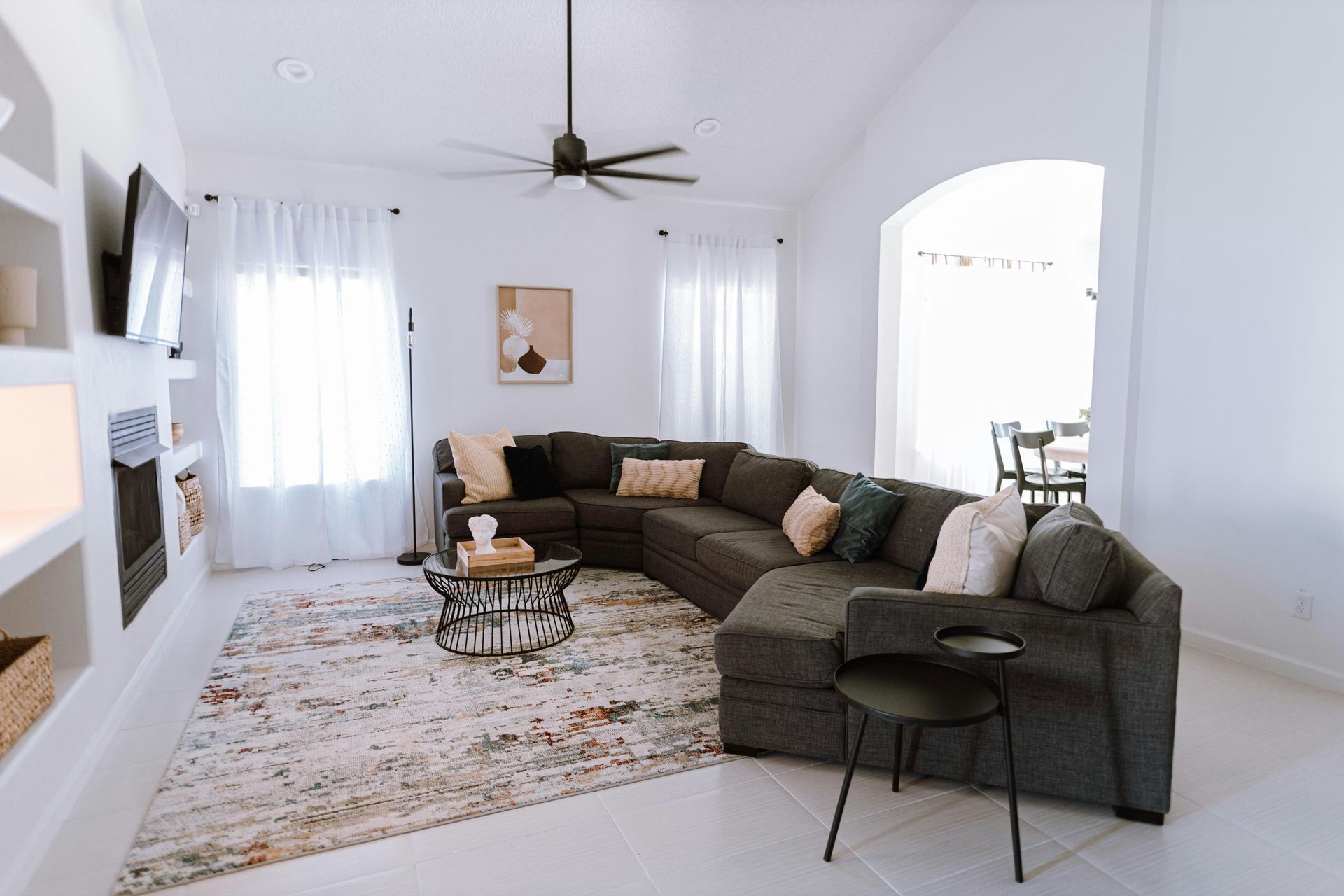 Living room with gray sectional sofa, rug, and built-in shelving. White walls, arched doorway, and ceiling fan.