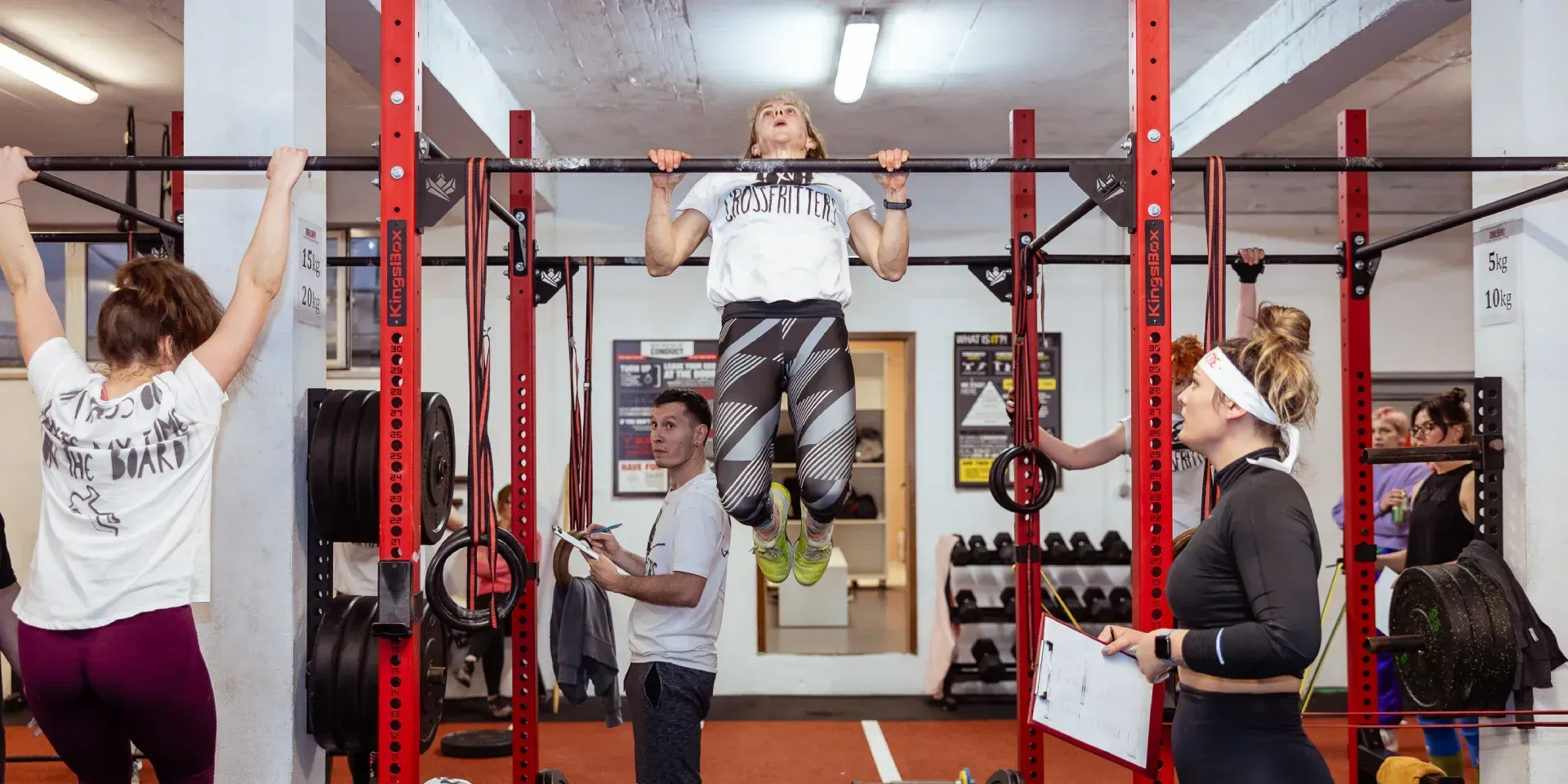 People exercising with weights in a gym. Some seated, some standing. A coach observes the workout.