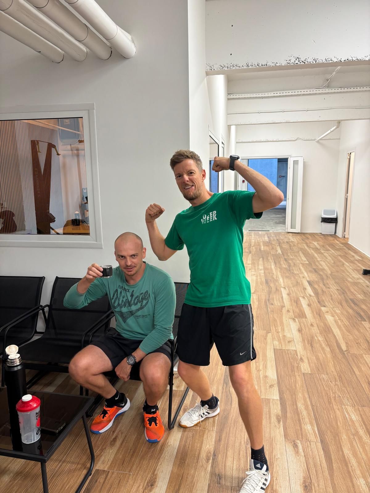 An older person performs a barbell squat in a gym. They wear a green hoodie and black pants.