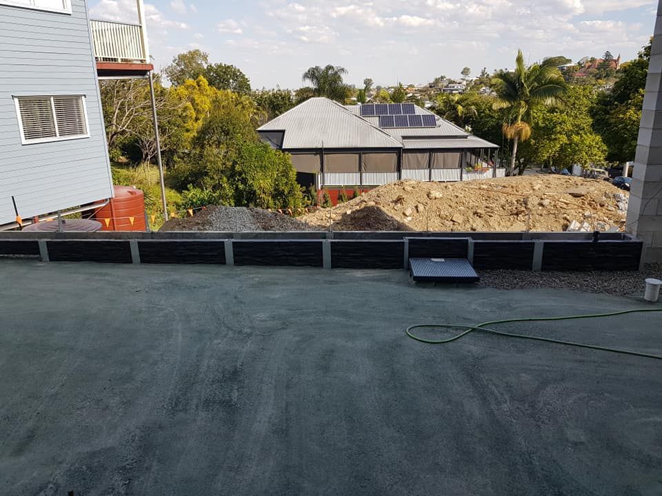 View of a construction site with a retaining wall, dirt pile, and houses in the background.