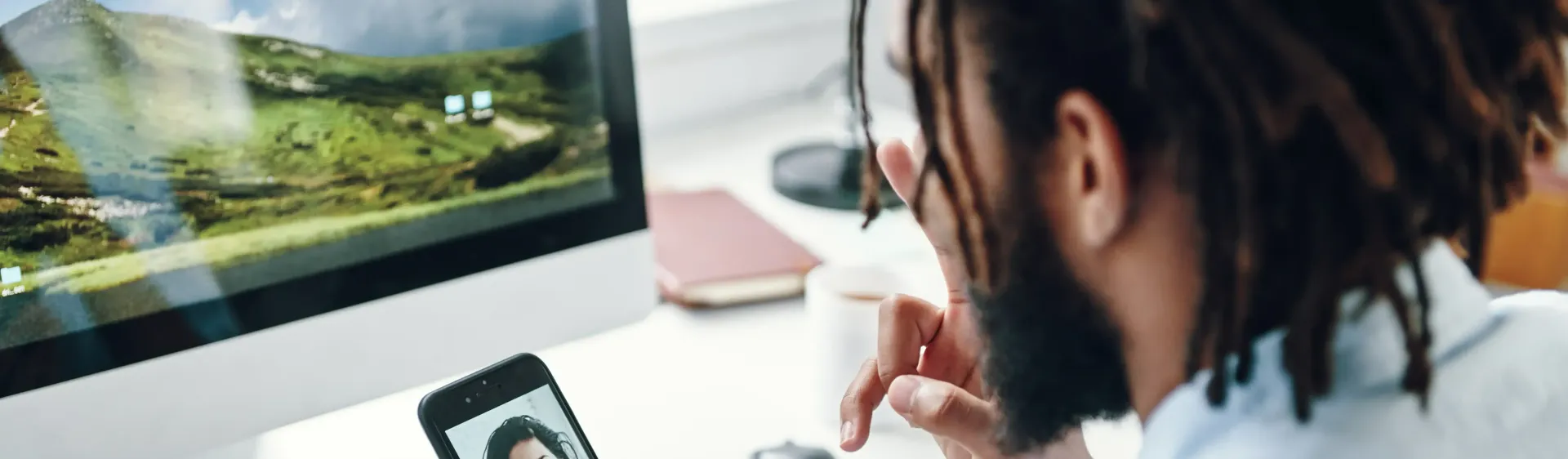 Man sitting at a desk in front of a computer using facetime on his phone.