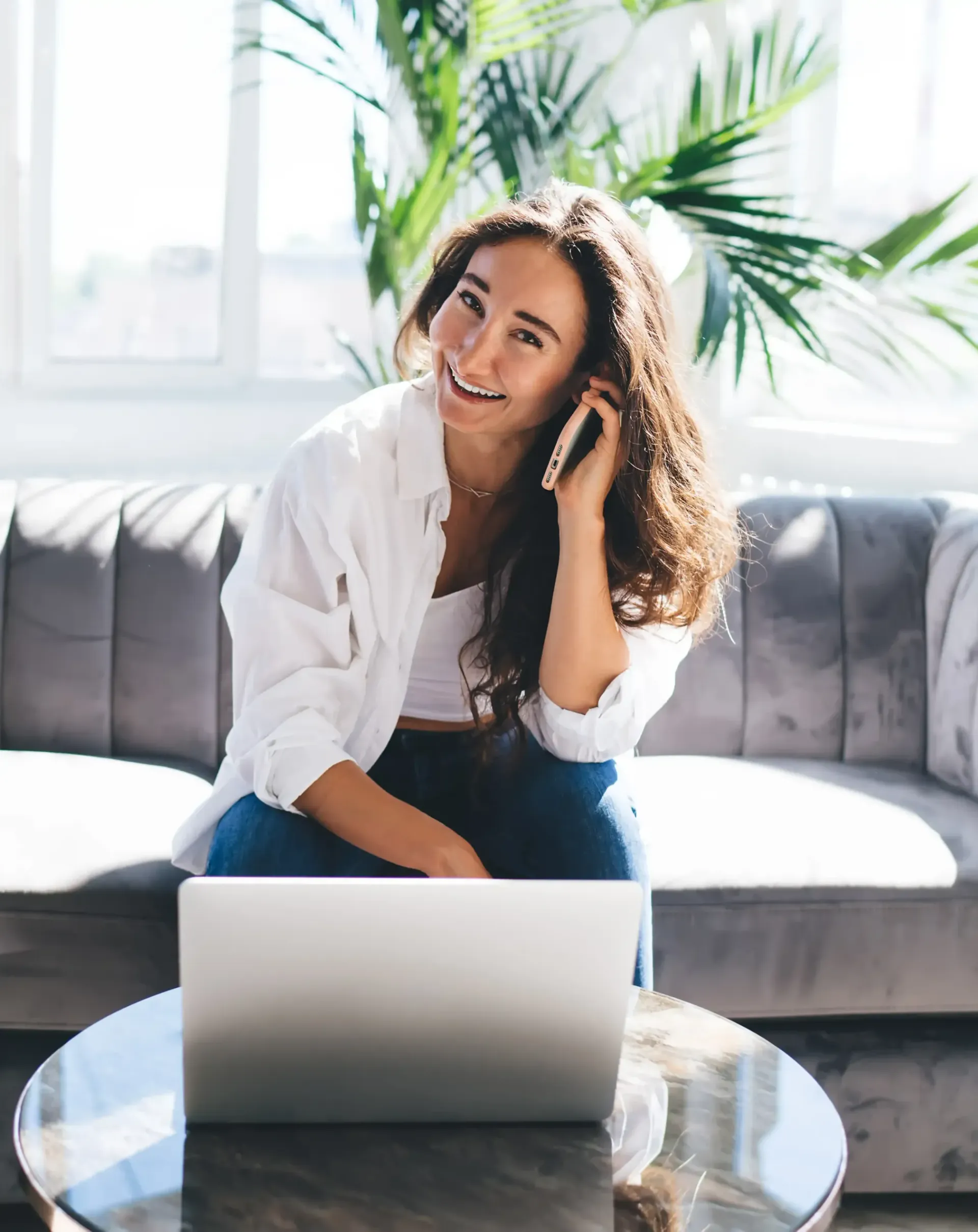 A woman is sitting on a couch talking on a cell phone doing cold outreach in front of a laptop computer.