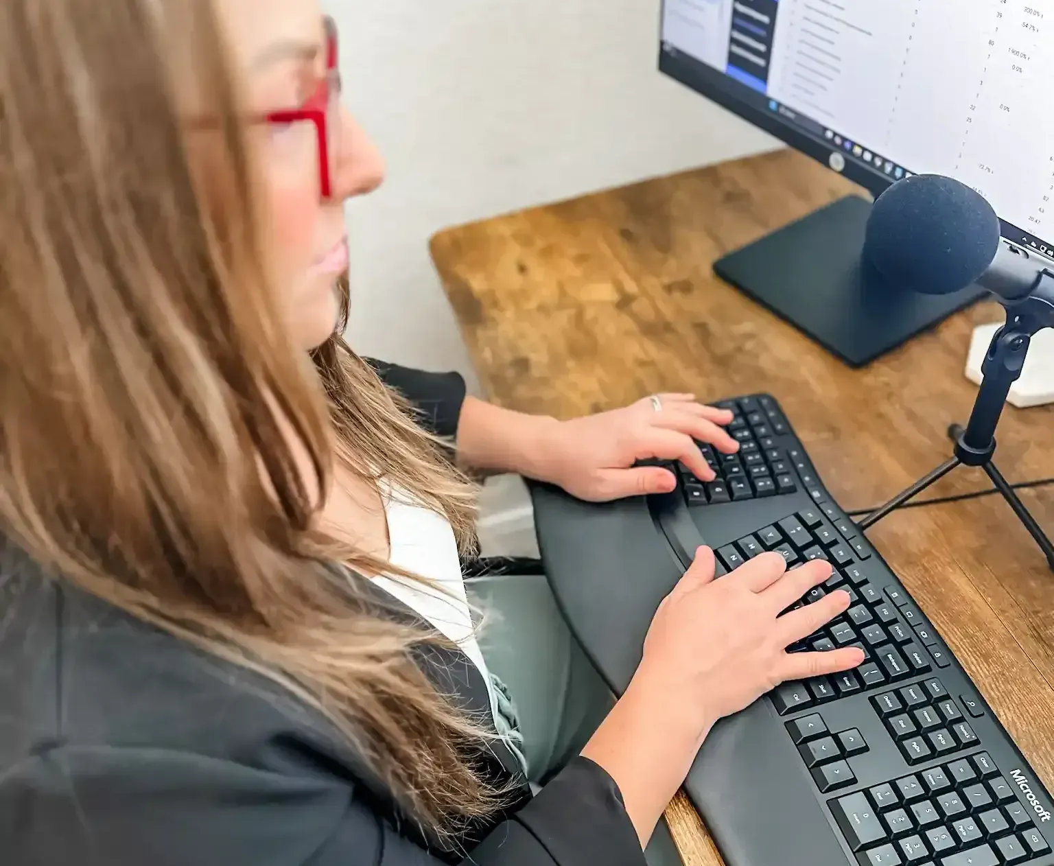 Amanda from AP Digital Service with red glasses types on an ergonomic keyboard teaching others about GEO, and optimized websites at a wooden desk, with a microphone and computer monitor visible.