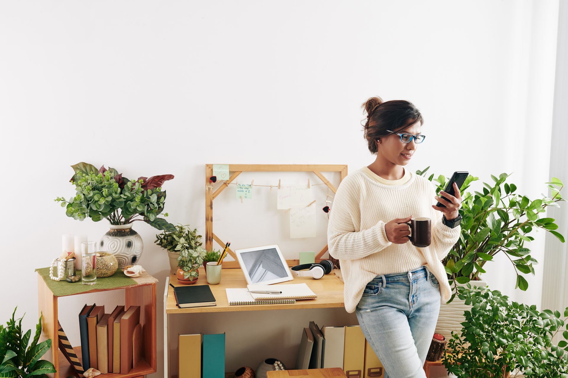 A woman is standing in front of a desk holding a cup of coffee and looking at her phone communicating with her coaching clients.