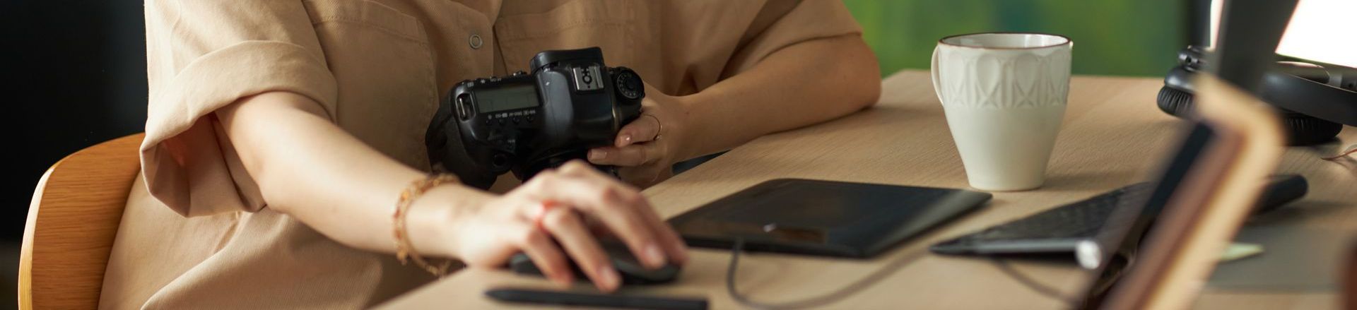 A woman is sitting at a desk holding a camera and using a computer mouse for her blogging revenue.