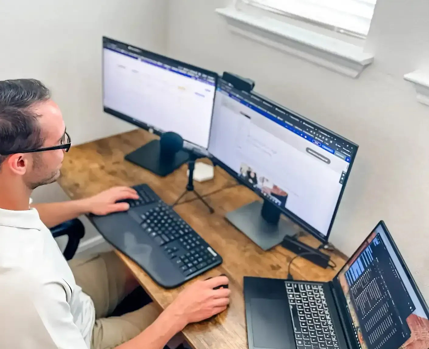 Peter from AP Digital Service at a desk working on a computer optimizing websites for GEO and SEO visibility with two monitors and a laptop. He's wearing glasses and a light-colored shirt.