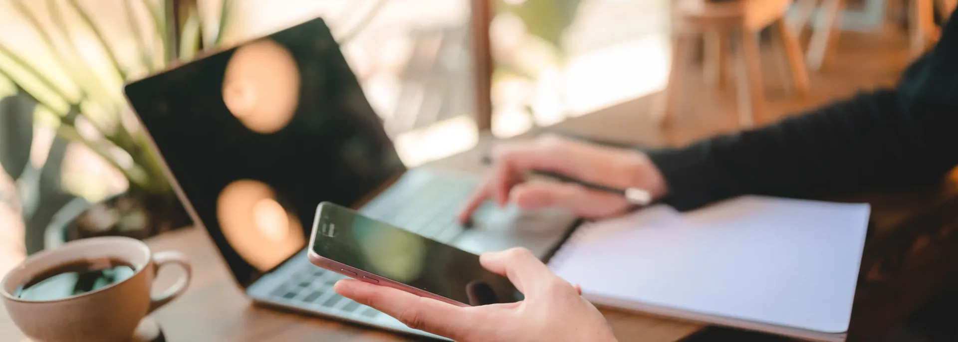 A person is sitting at a table using a laptop and a cell phone to bring in clients to their coaching business using SEO.