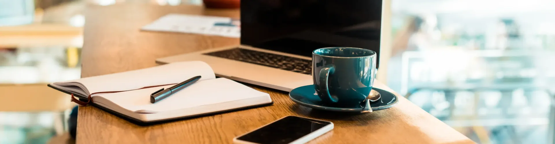 A laptop , notebook , cell phone , and cup of coffee are on a wooden table ready to be utilized for SEO and AI visibility for Coaches and Creators.