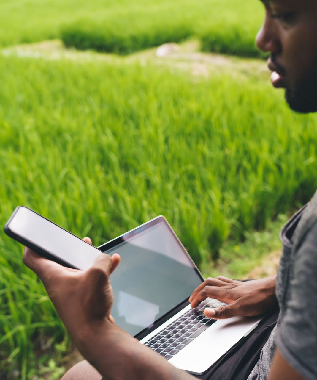 A man is sitting in a field using a laptop and a phone to view his evergreen content.