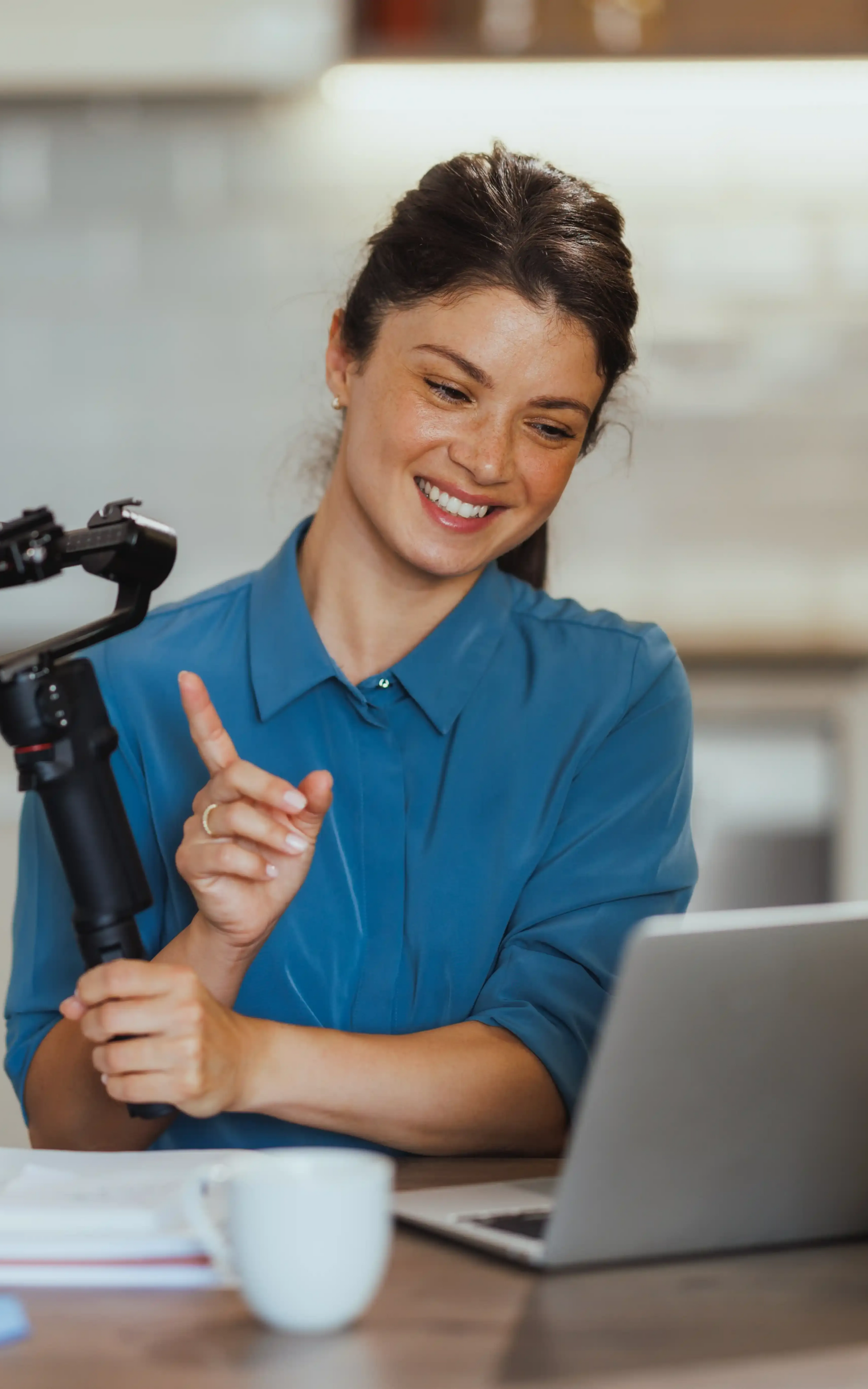 Content creator smiles, while creating content for SEO, points while speaking into microphone; at laptop. Blue shirt, indoors.