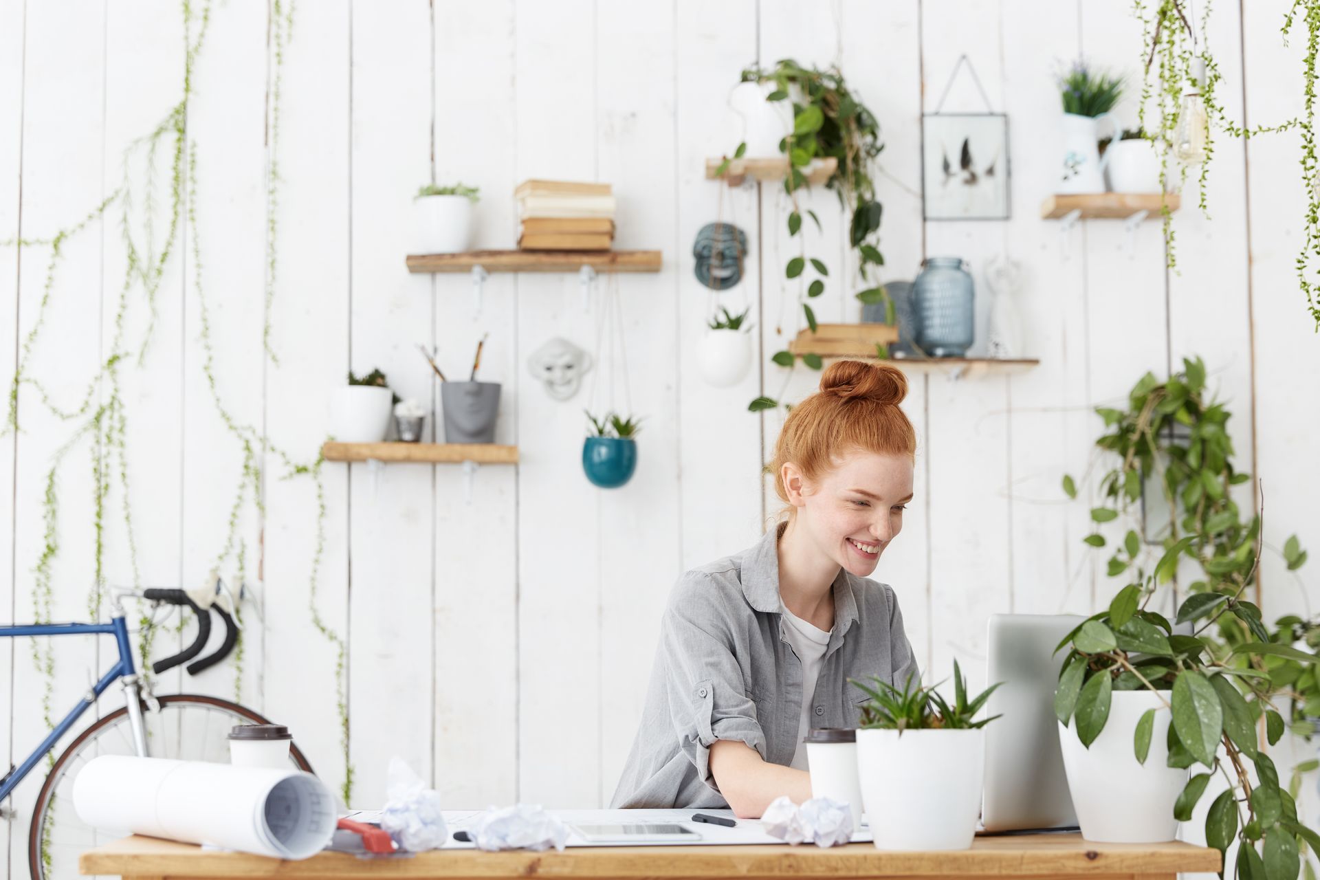 A woman is sitting at a desk using a laptop computer surrounded by potted plants while coaching her clients.