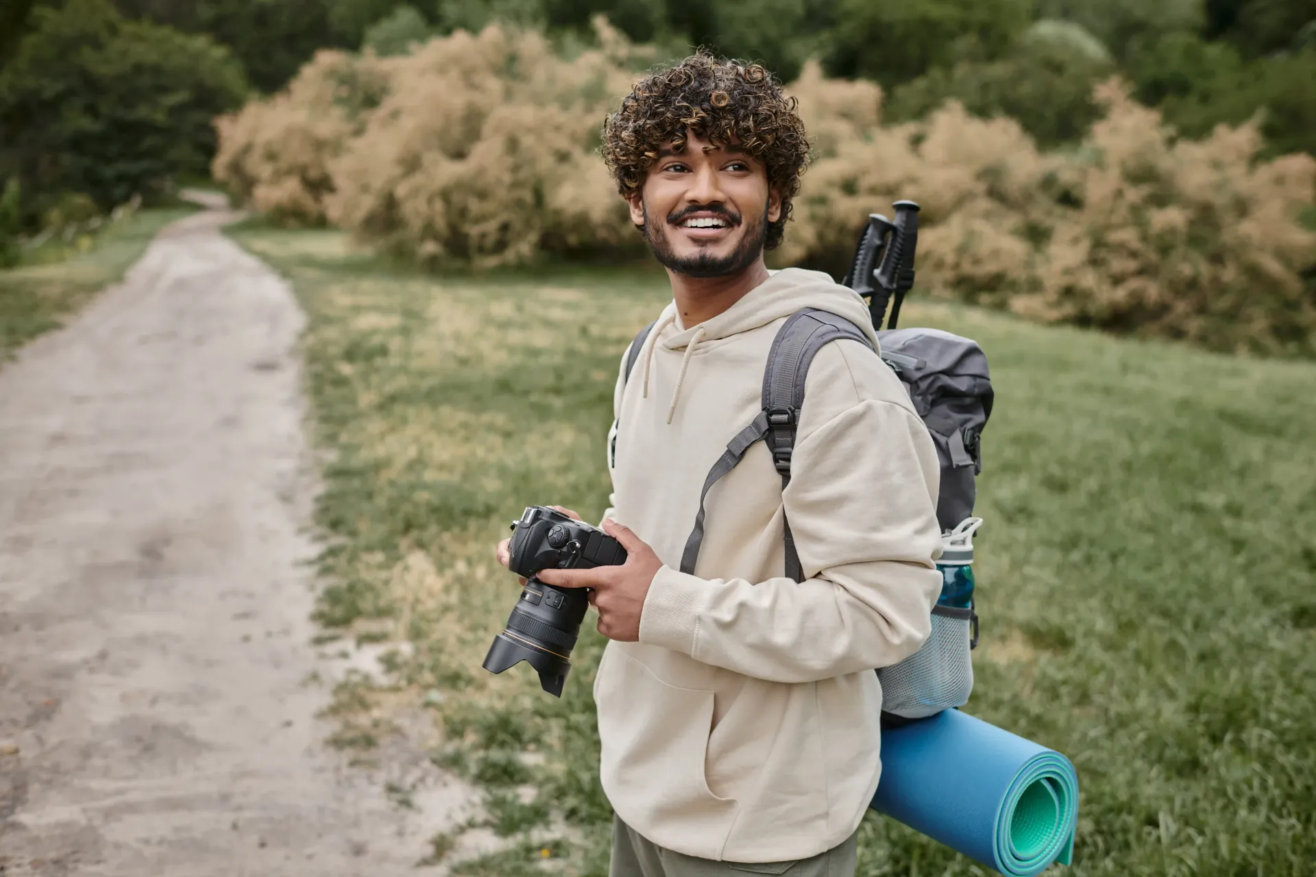 A man with a backpack is holding a camera and a yoga mat.