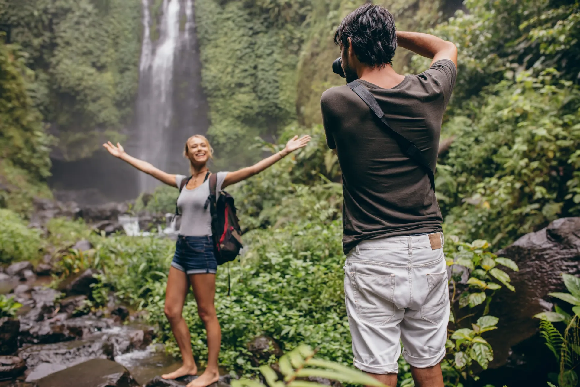 A man is taking a picture of a woman in front of a waterfall.