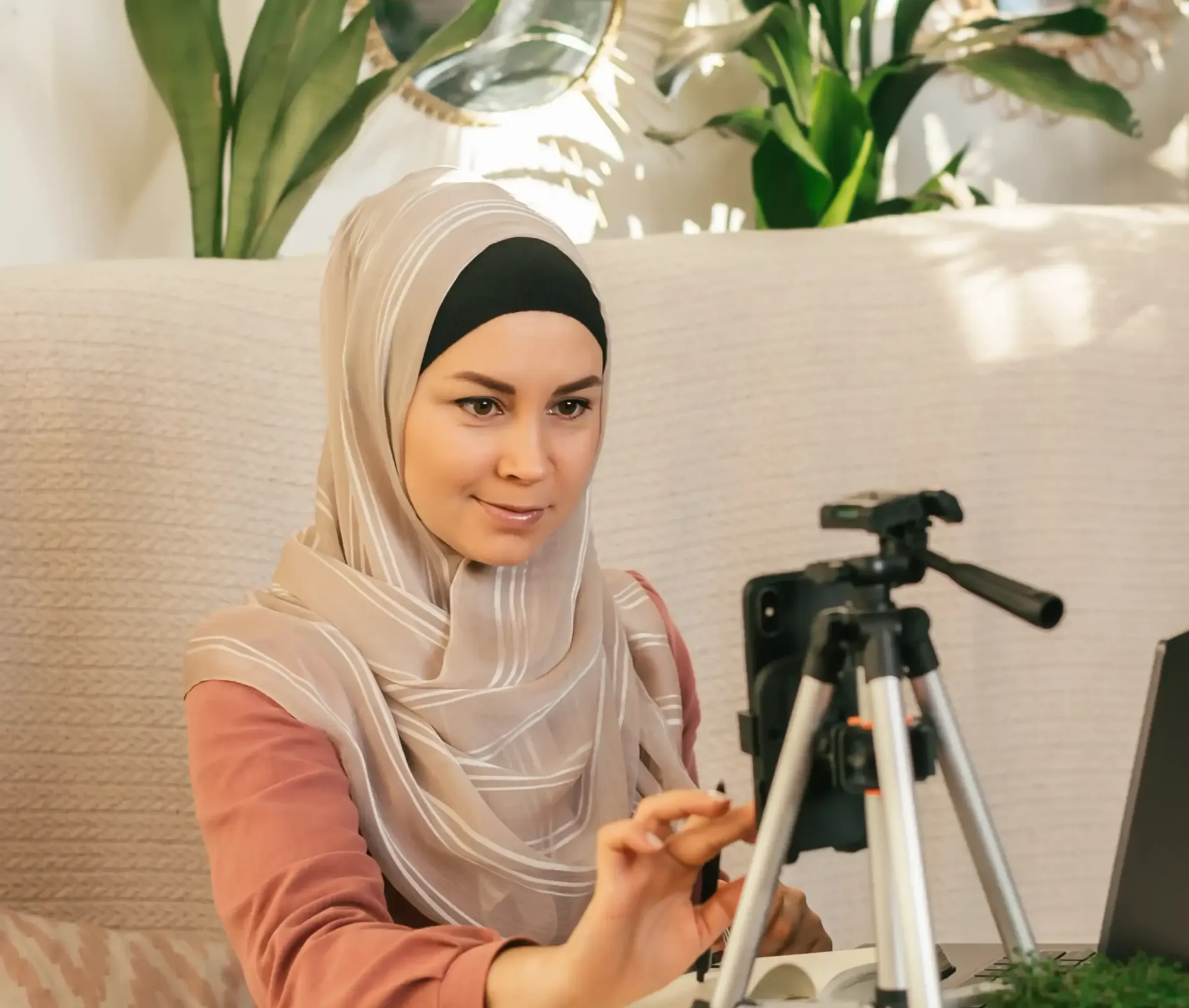 A woman in a hijab is sitting in front of a laptop and a camera on a tripod