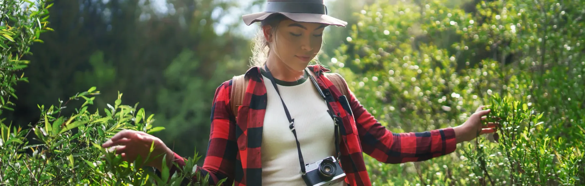 A woman in a plaid shirt and hat is walking through a forest.
