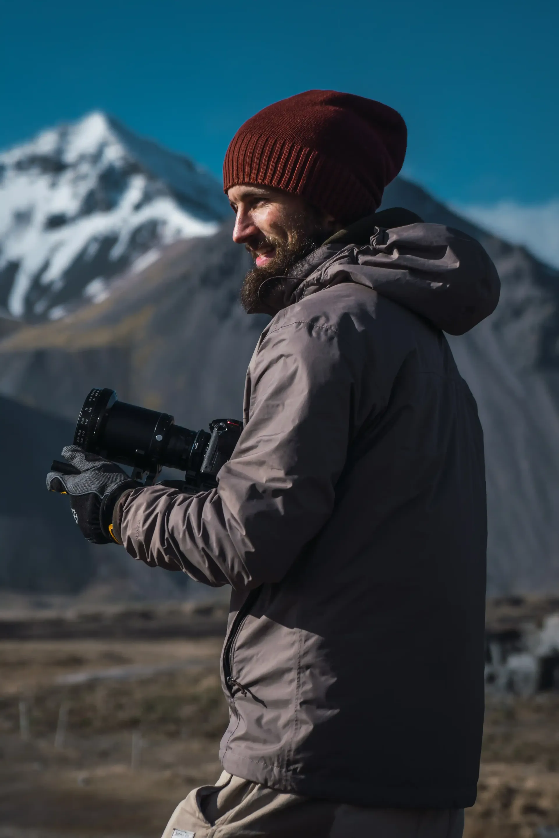 A man with a beard is holding a camera in front of a mountain.