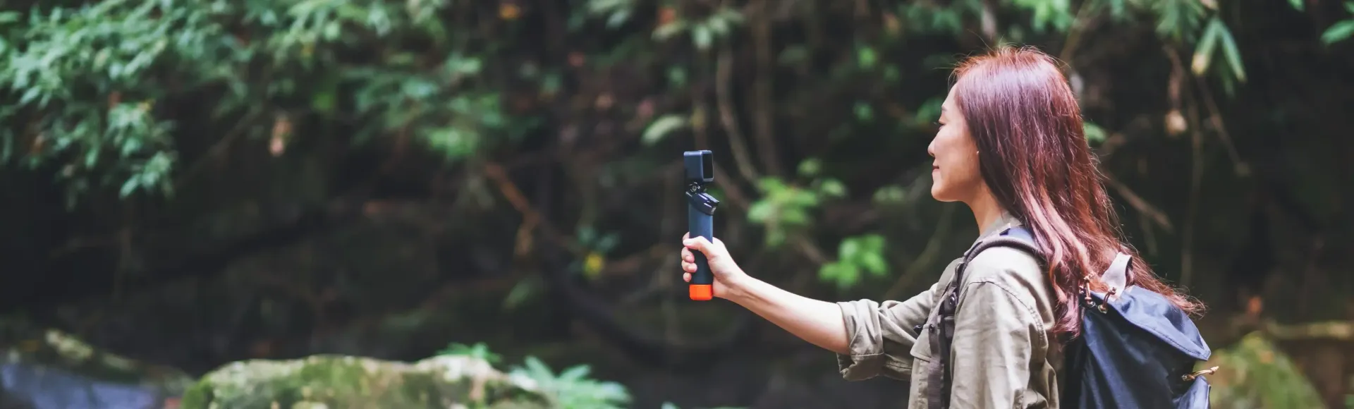 A woman with a backpack is taking a picture of a waterfall with her cell phone.