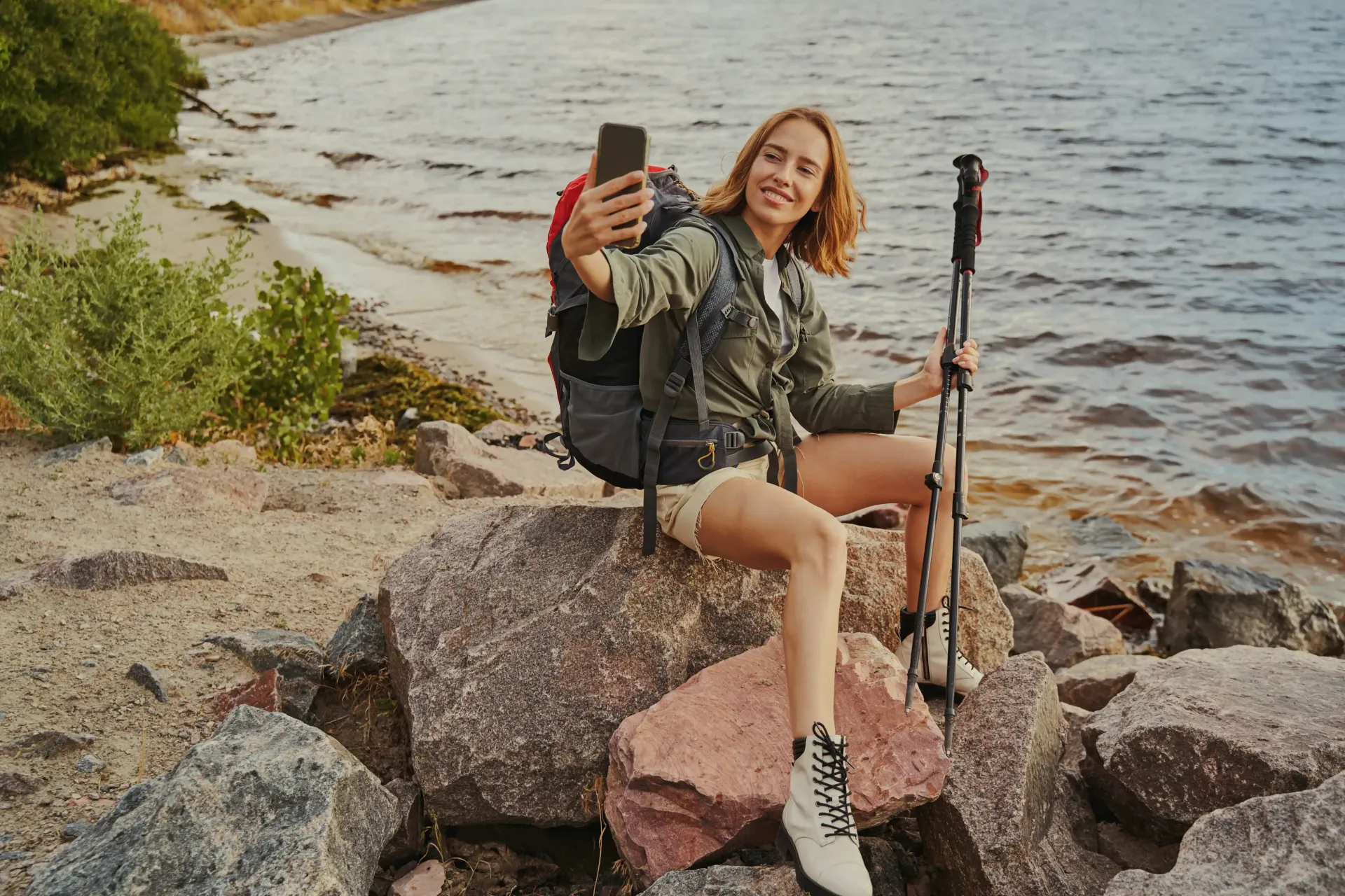 A woman with a backpack is sitting on a rock taking a selfie.