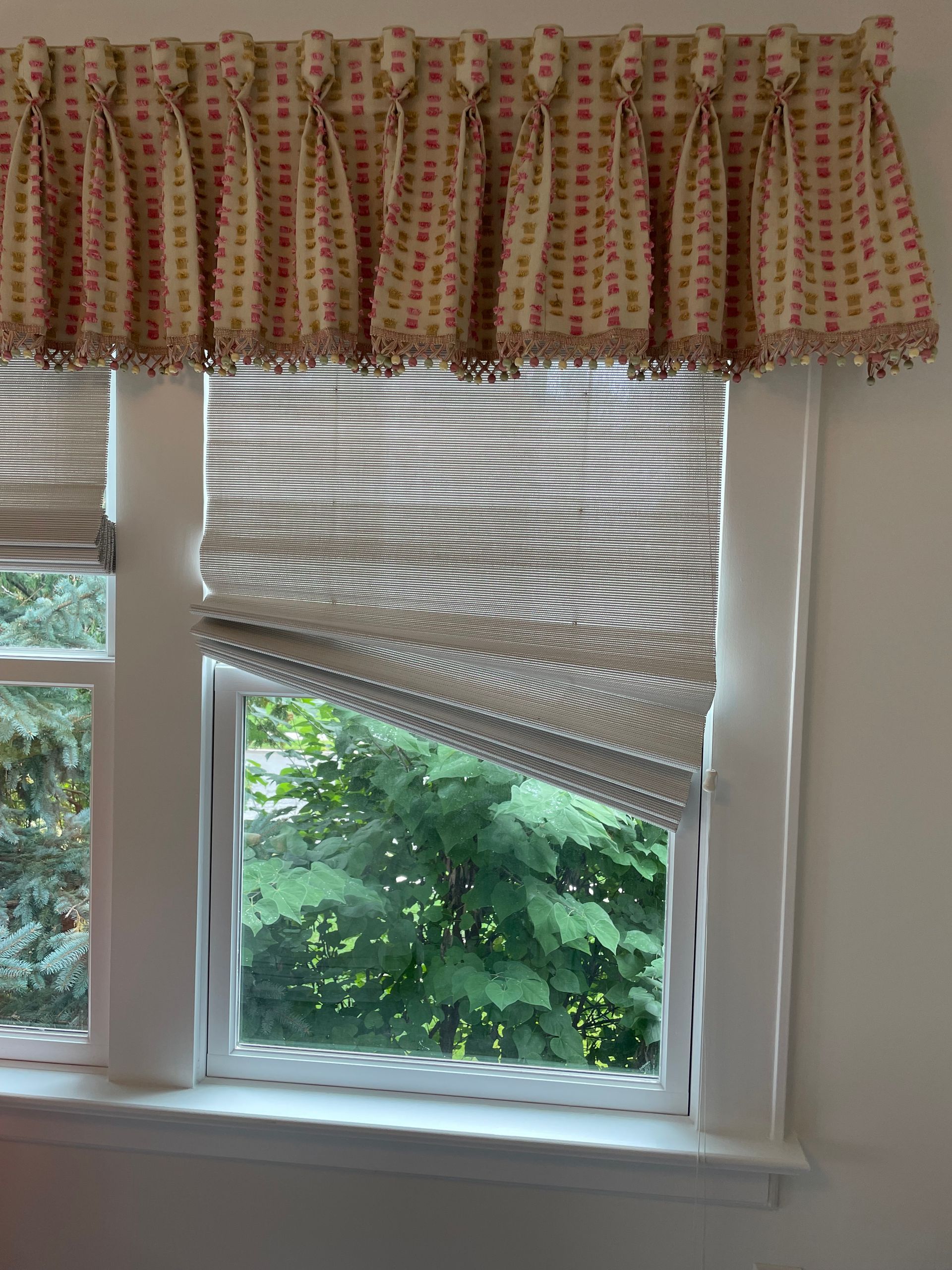 Window with a patterned valance and a striped roman shade, overlooking lush greenery.