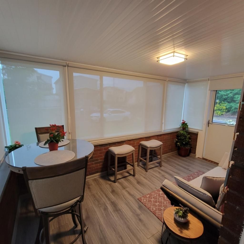 Sunroom with a table, stools, couch, and potted plants. The room has large windows covered with roller shades.
