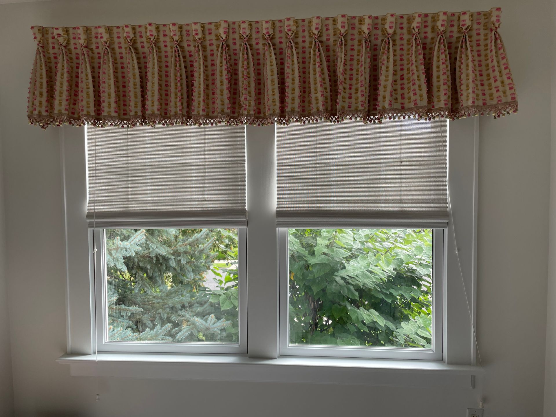 Window with two panes, each covered by a beige shade. A valance with red pattern tops the window. Green trees are visible.