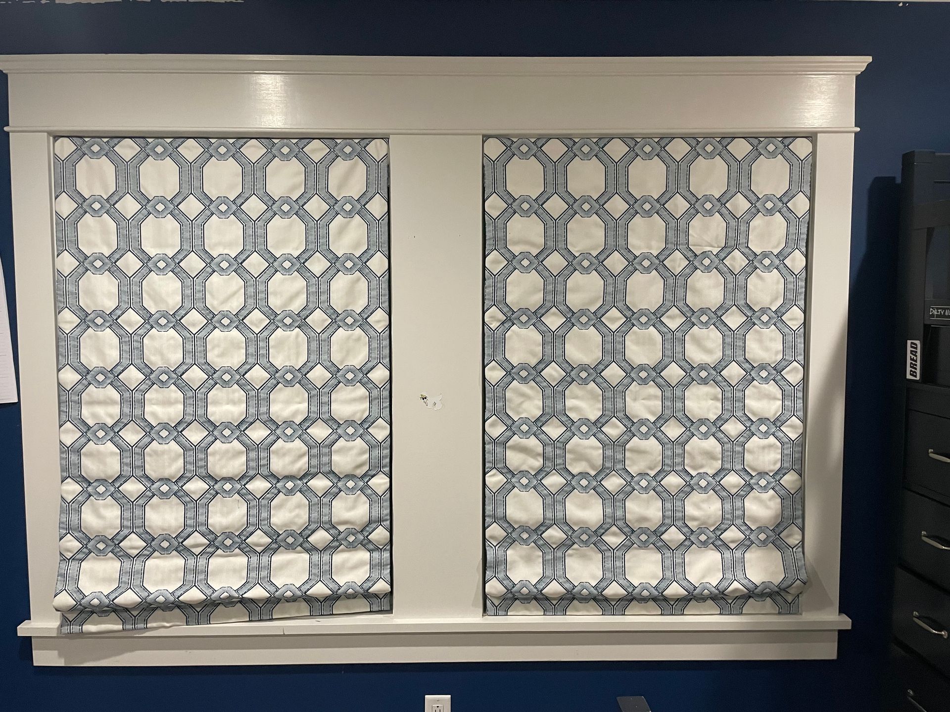 Two white-framed windows with matching blue and white geometric patterned shades, set against a dark blue wall.