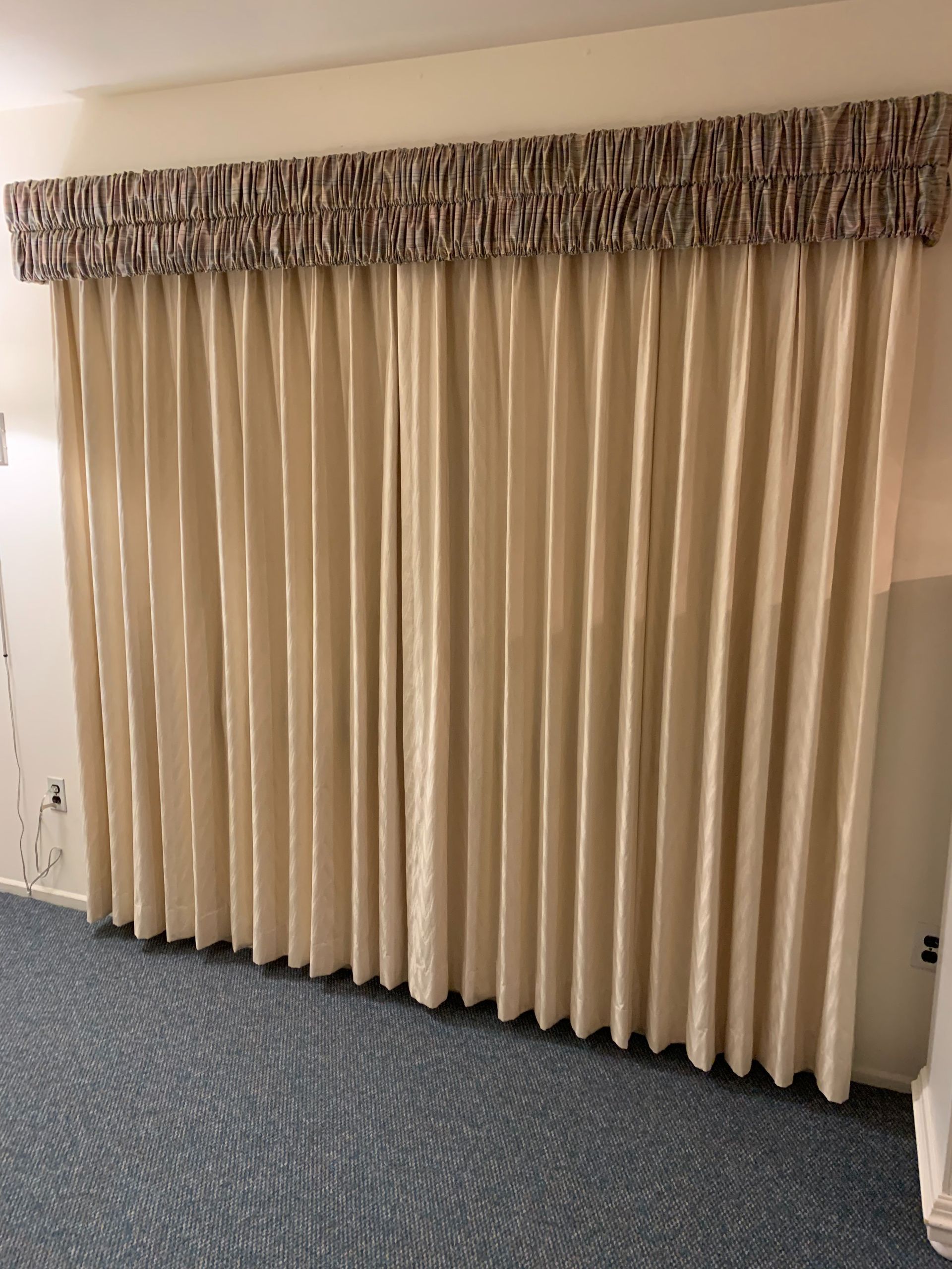 Beige pleated curtains with a patterned valance, hanging over a blue patterned carpet.