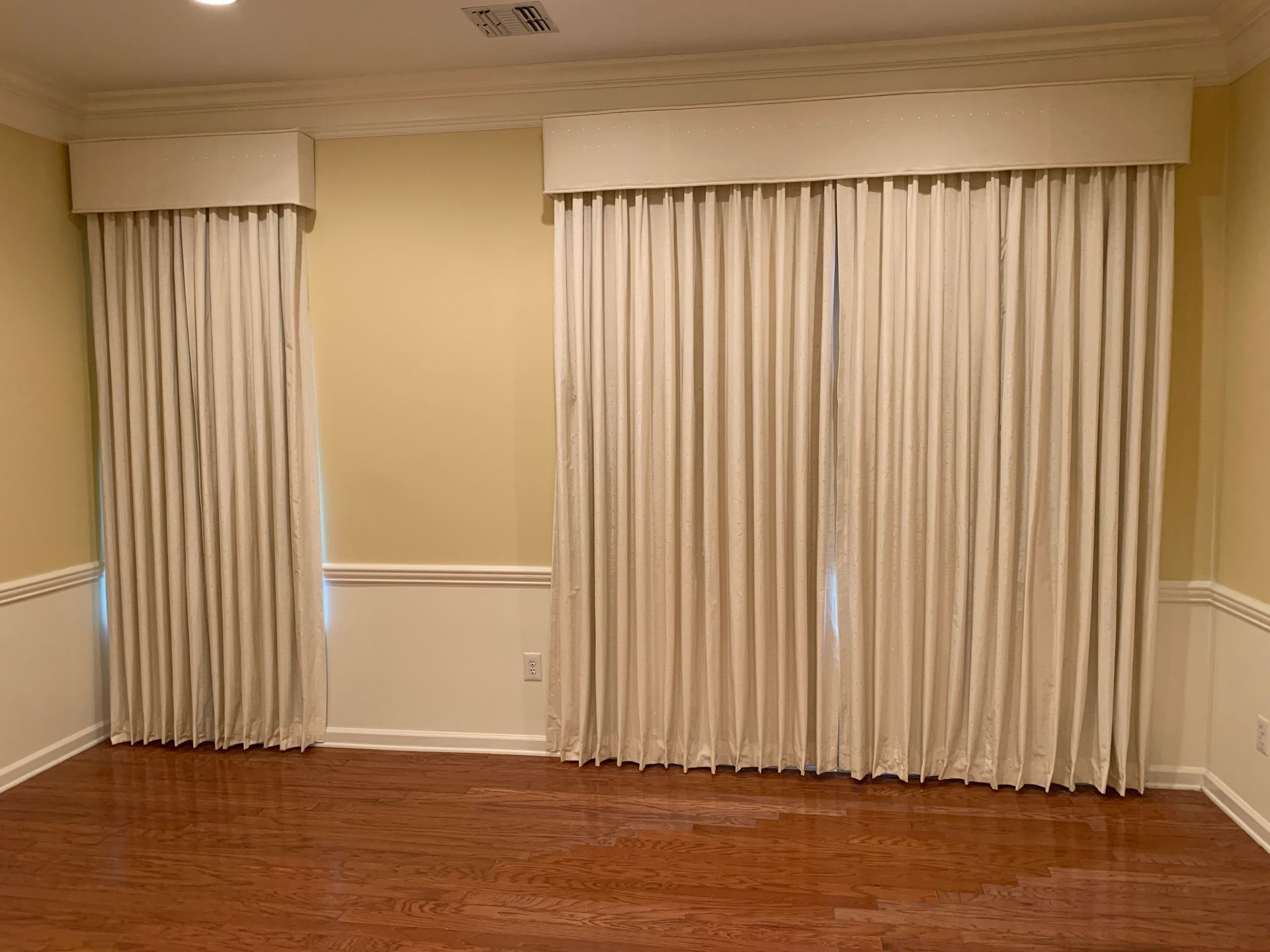 Cream curtains hang against a light yellow wall in a room with a wood floor.