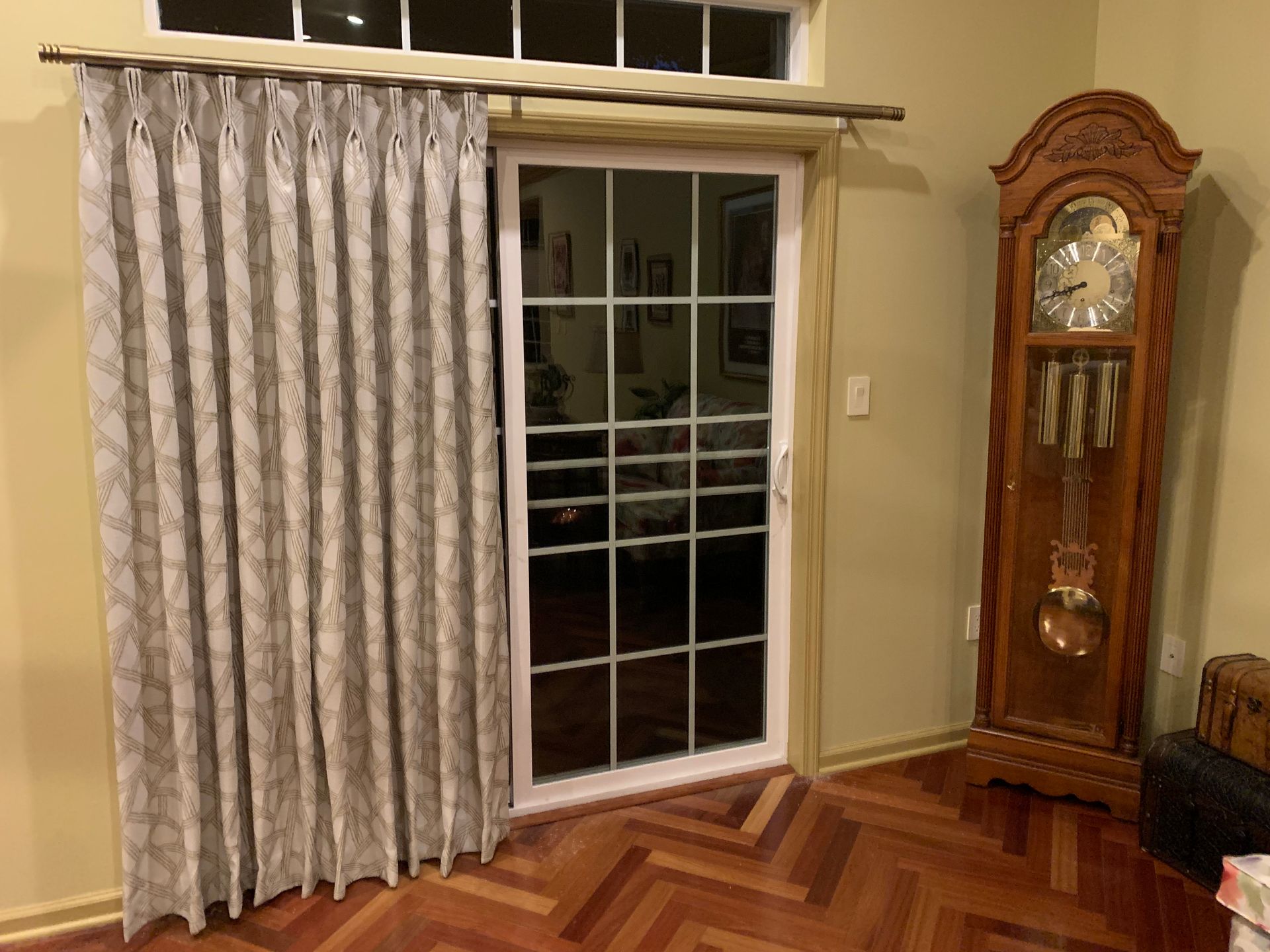 Sliding glass door with patterned curtain partially closed, near a grandfather clock. Wooden floor and cream-colored walls.