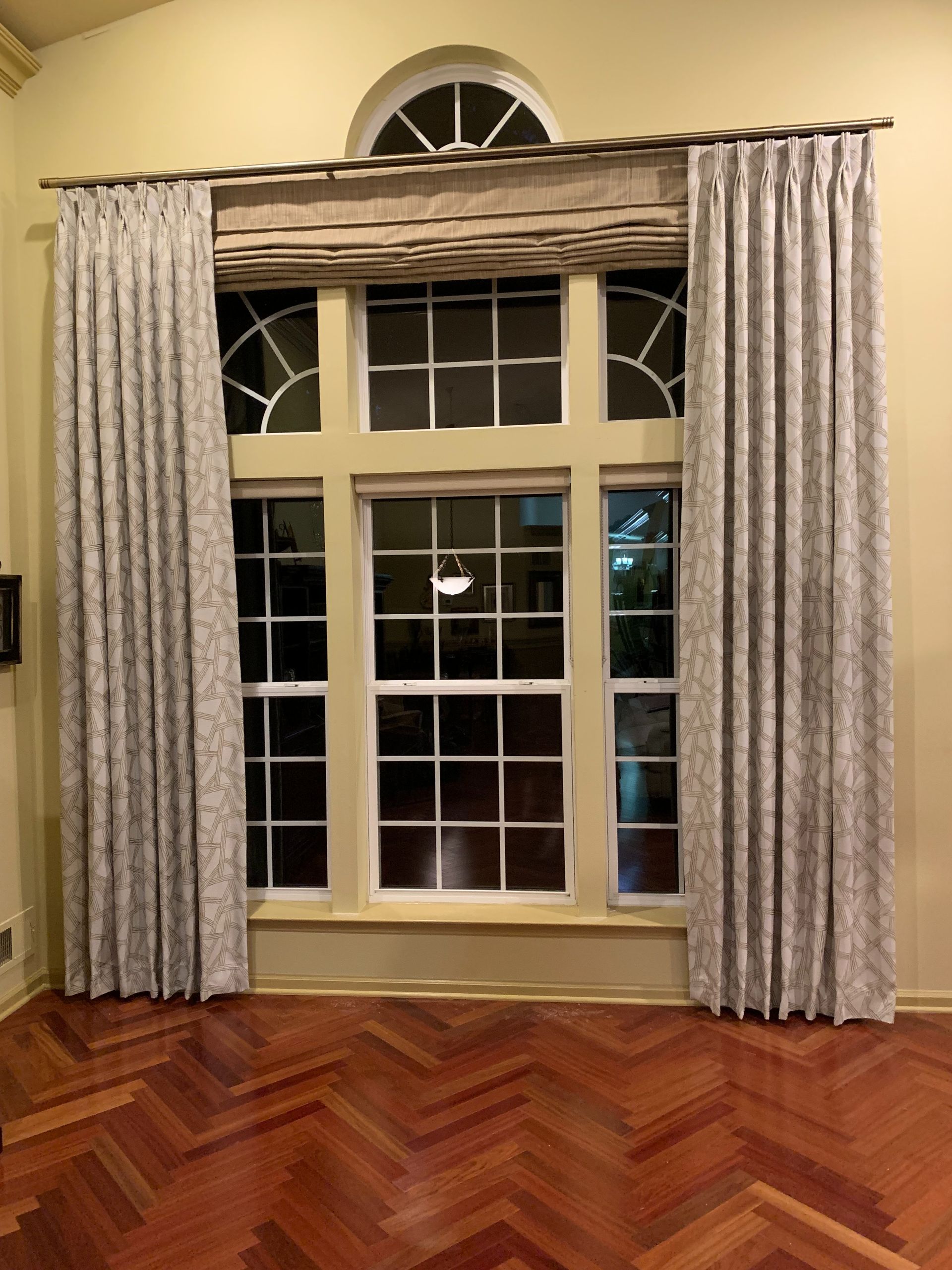 Window with curtains and a Roman shade above a hardwood floor.