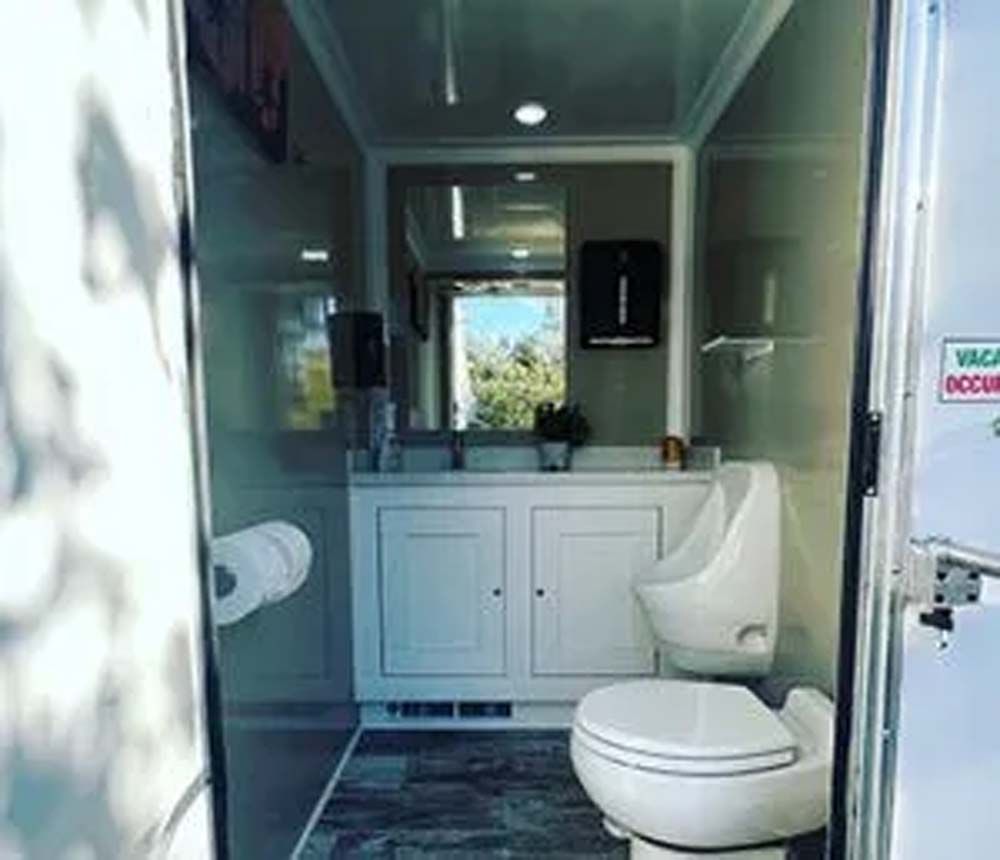 Interior of a luxury portable restroom. White cabinets, two sinks, urinal, toilet, and a mirror.